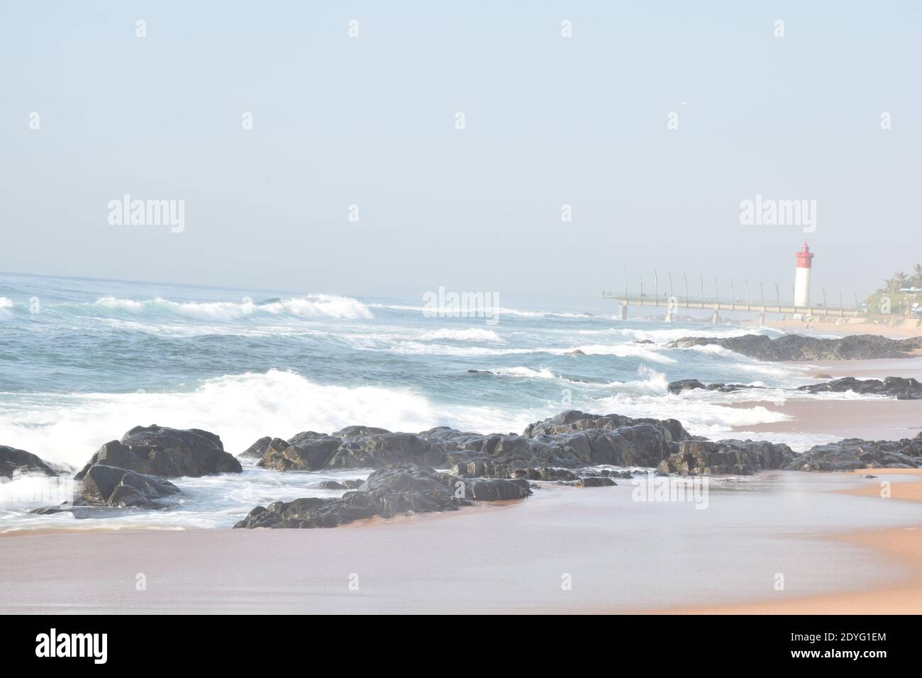 La mattina presto sulla spiaggia, le onde si schiantano contro le rocce con il molo e il faro sullo sfondo Foto Stock