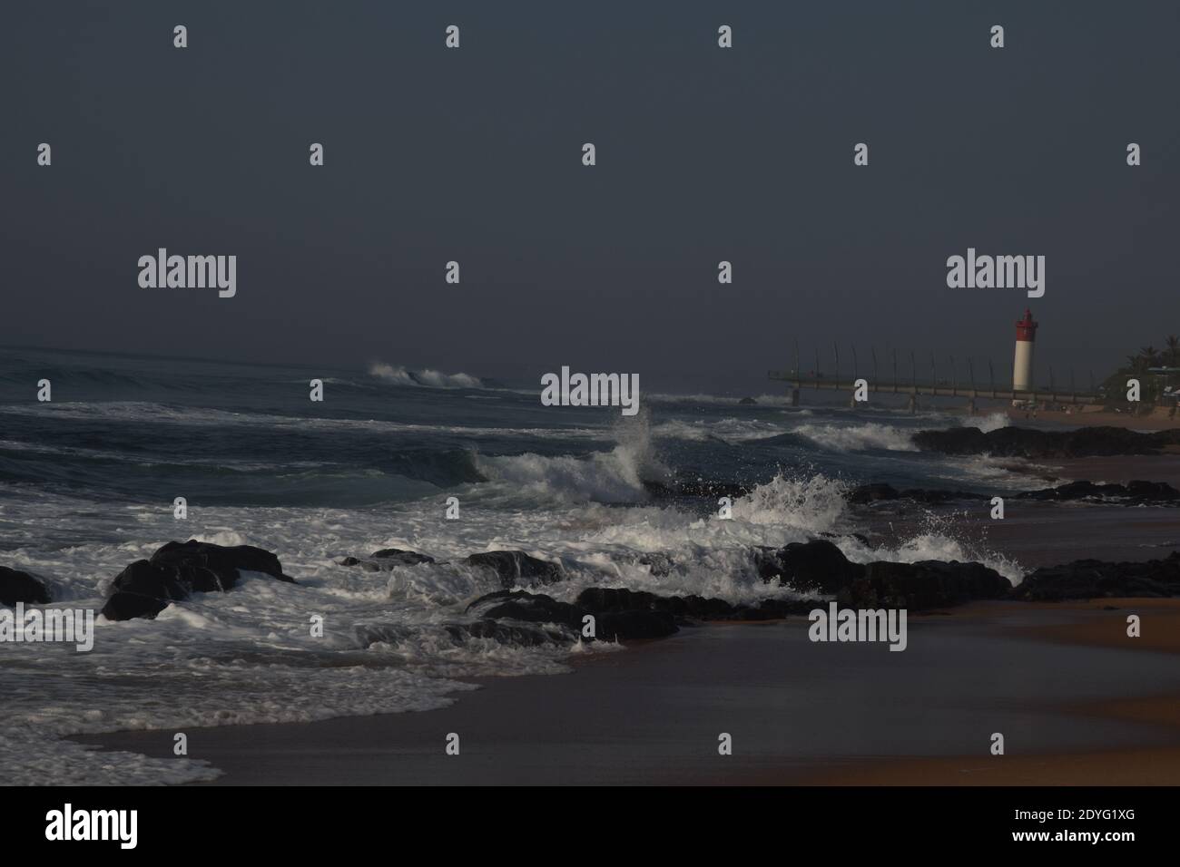 La mattina presto sulla spiaggia, le onde si schiantano contro le rocce con il molo e il faro sullo sfondo Foto Stock