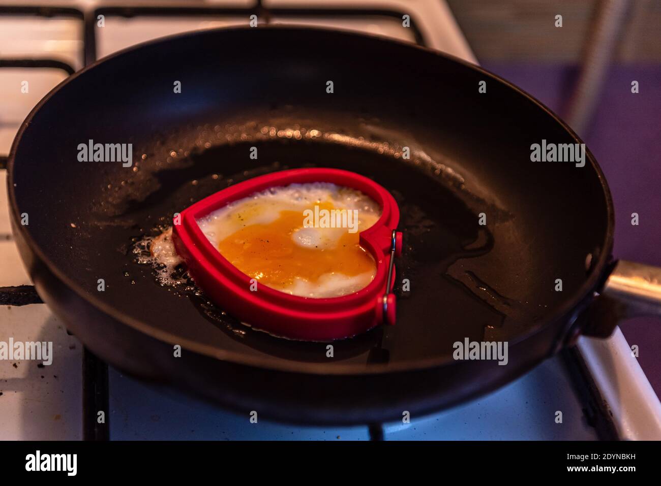 uovo fritto in padella a forma di cuore su una stufa a gas bianca e griglia nera, che tiene la padella sopra una fiamma fuoco Foto Stock