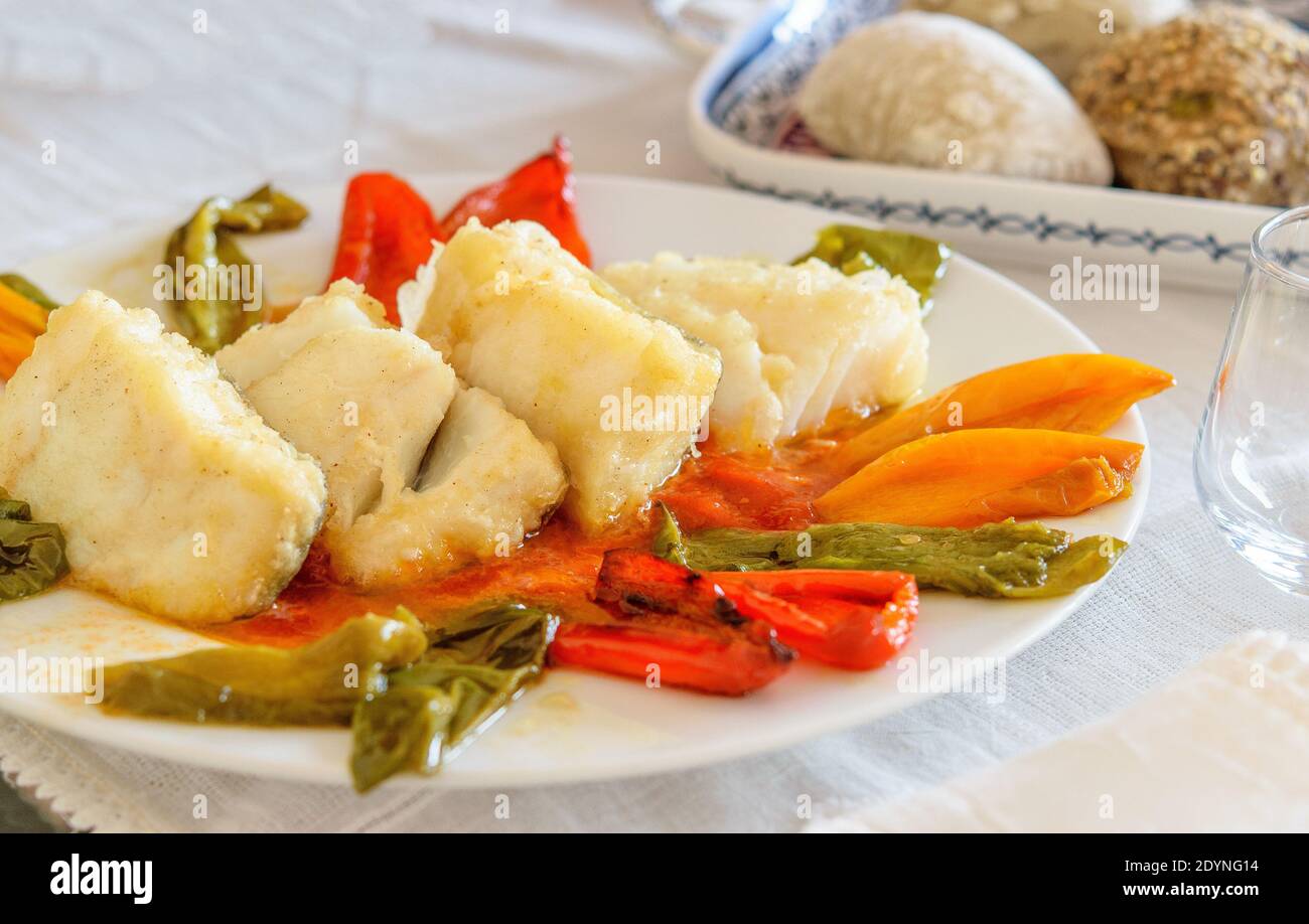 Fette di pesce di merluzzo caldo fritto con fette di peperoni dolci sul piatto bianco. Un piatto con ciambelle sul tavolo servito. Pesce sano di mediterran Foto Stock