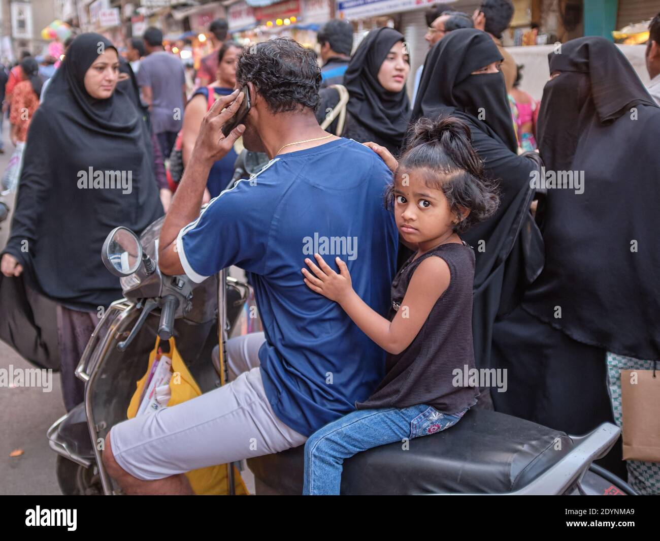 Circondato da donne musulmane in burqas, un padre ferma la sua moto per fare una telefonata, sua figlia aggrappato a lui sulla sede posteriore; Mumbai, India Foto Stock