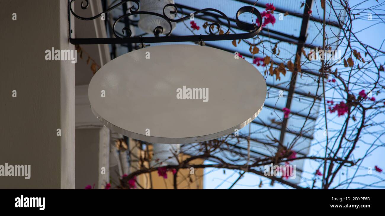 Cartello ovale in metallo bianco vuoto, mockup commerciale, dima. Ritaglio vuoto del cartellone appeso alla base in metallo nero all'esterno del negozio, copyspace. Sfocatura Foto Stock