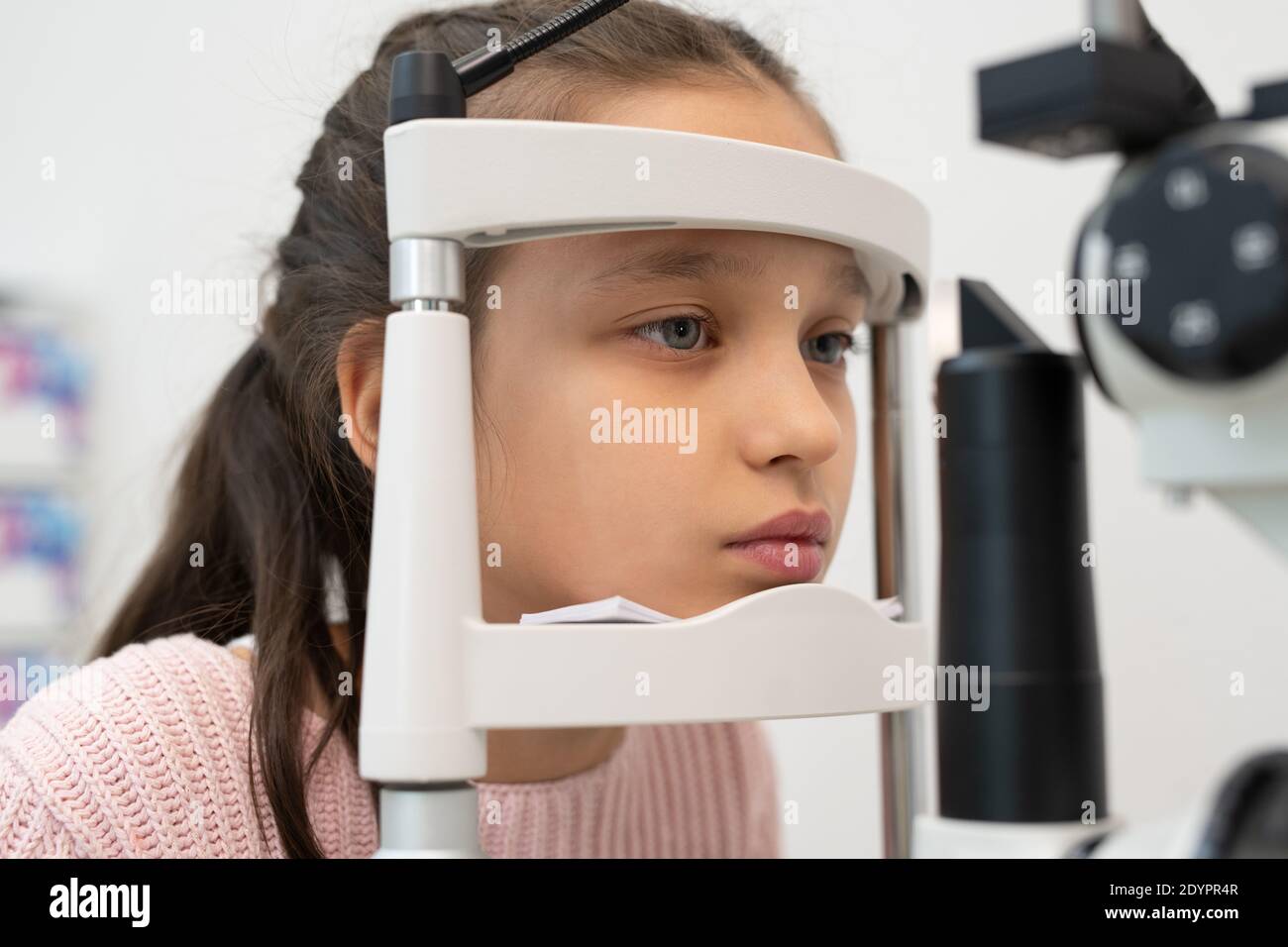 Volto di scolastica elementare seduto di fronte ad apparecchiature oftalmologiche mentre la sua vista ha controllato nelle cliniche moderne Foto Stock