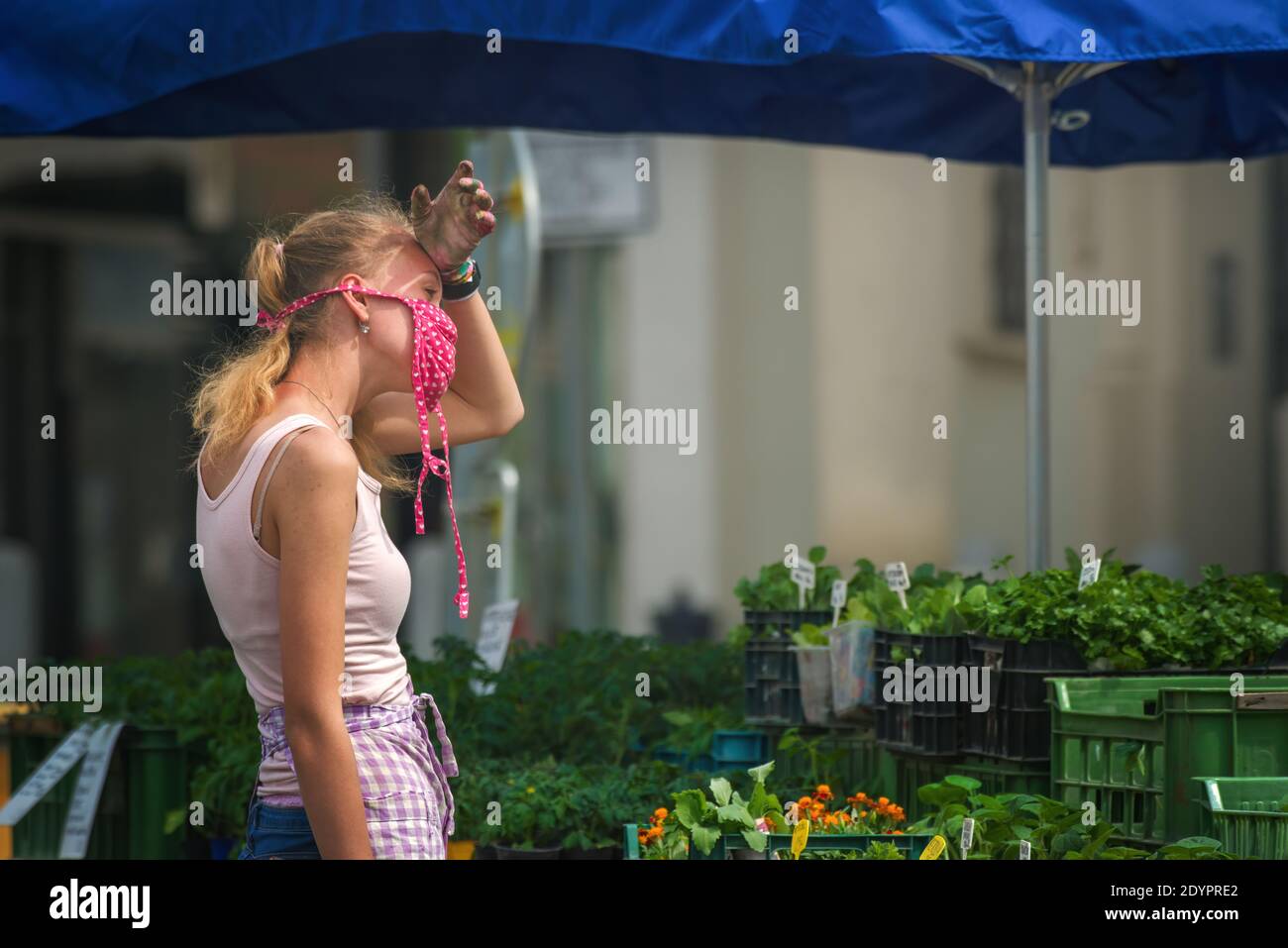 Stanco venditore che indossa una maschera facciale in tessuto ad un mercato agricolo che ha un mal di testa dal clima clammy. Foto Stock