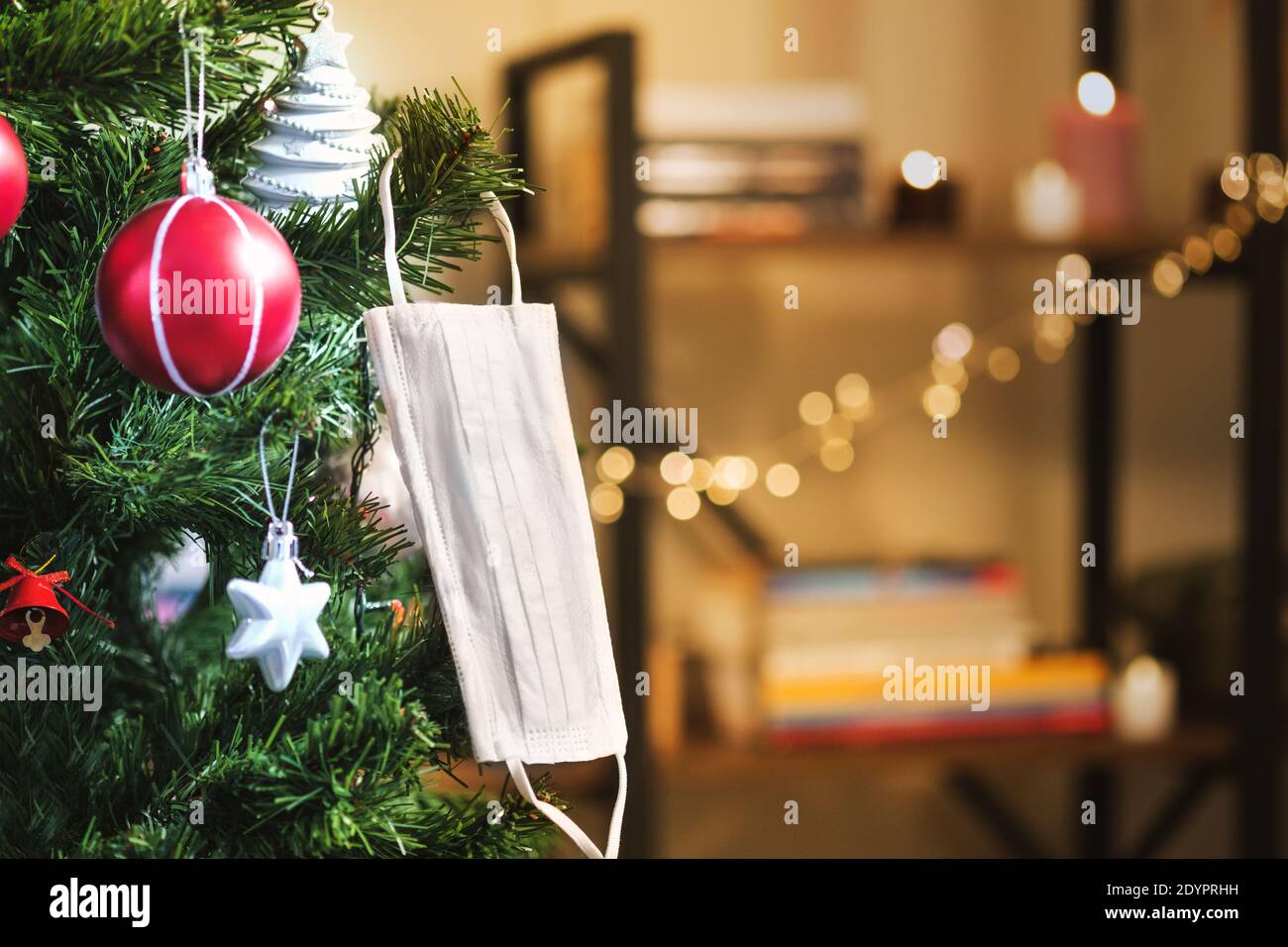 Ornamenti di Natale e una maschera medica che pende su un ramo di un albero di natale in una stanza con una mensola di libro sfocata sullo sfondo. Foto Stock