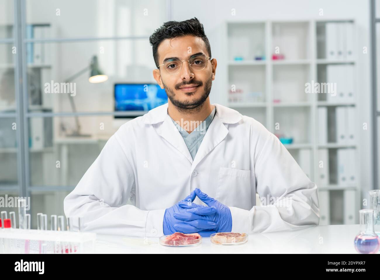 Giovane ricercatore scientifico di successo in whitecoat che ti guarda mentre seduto sul posto di lavoro con campioni di carne vegetale grezza in capsule di petri Foto Stock