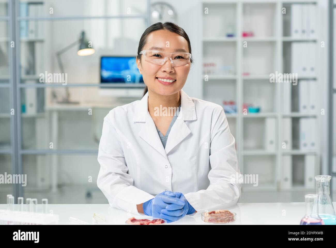 Felice giovane donna asiatica guanto in bianco e occhiali seduta sul posto di lavoro con due campioni di carne vegetale cruda in capsule di petri Foto Stock