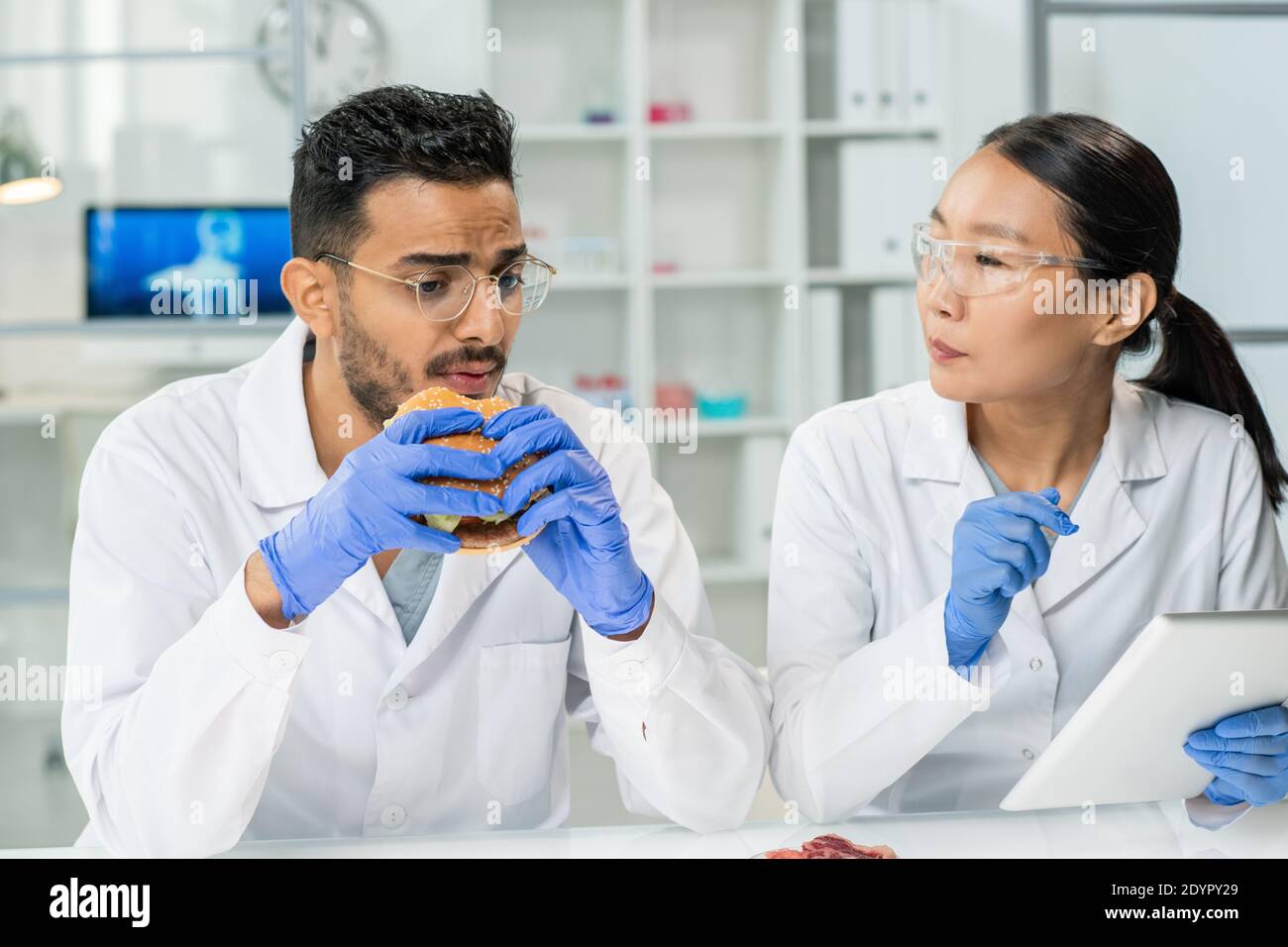 Gustato giovane lavoratore maschio di controllo alimentare mangiando hamburger da luogo di lavoro e descrivendo il suo gusto alla collega femminile con tablet in laboratorio Foto Stock
