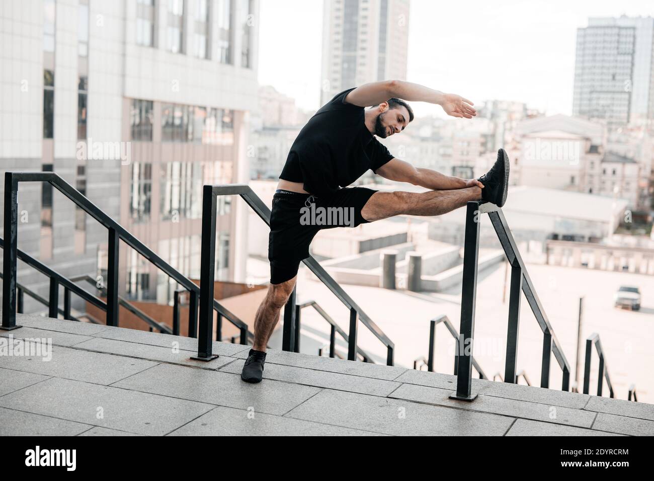 Uomo forte che si riscalda. Attività mattutina. Uomo che stende le braccia e si guarda via. Allenamento mattutino con corrimano. Abbigliamento sportivo nero. Fit uomo facendo Foto Stock