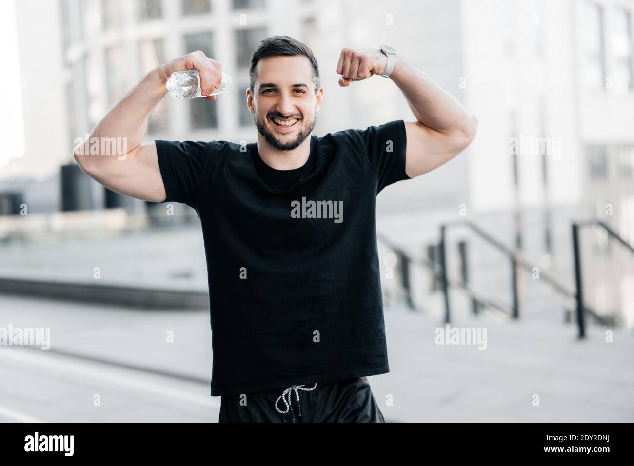 Uomo allegro che mostra i suoi muscoli. Concetto di allenamento duro. Fit uomo che mostra bicipite e sorridente. Grigio mattina città sullo sfondo. Bell'uomo ha excercised a. Foto Stock