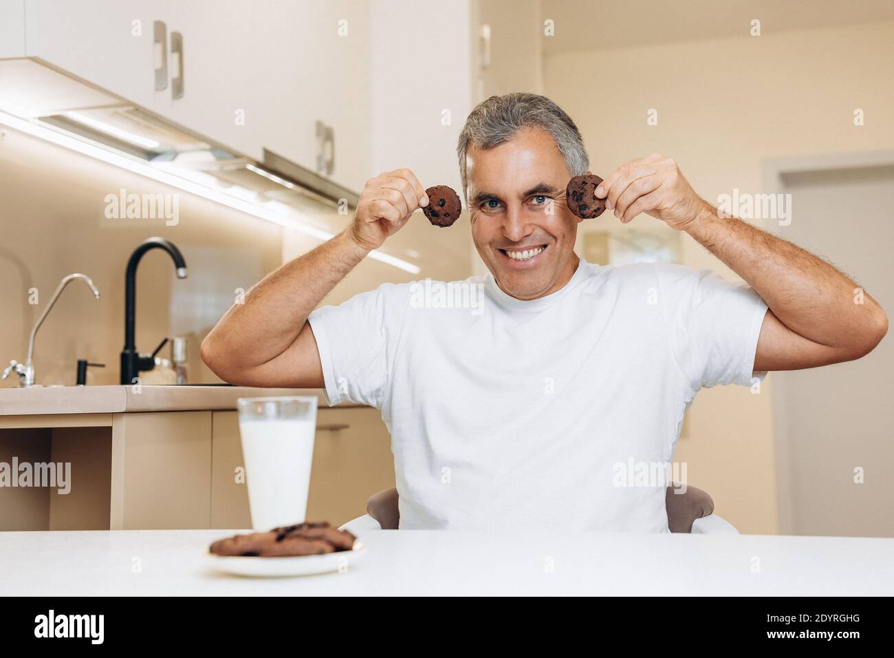 Uomo anziano divertente che tiene biscotti vicino al viso e sorridente con i denti. Concetto di volti pazzi. Uomo che si aggira e cerca di far ridere la sua famiglia Foto Stock