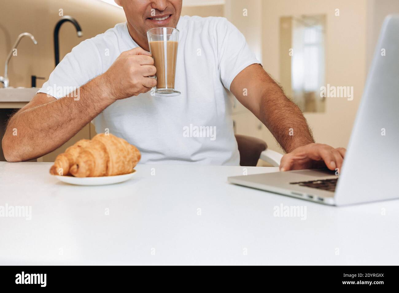 L'uomo soddisfatto beve latte o caffè con latte e usa il suo computer portatile moderno. Croissant croccante su un tavolo bianco. Uomo che guarda video divertenti e si diverti Foto Stock