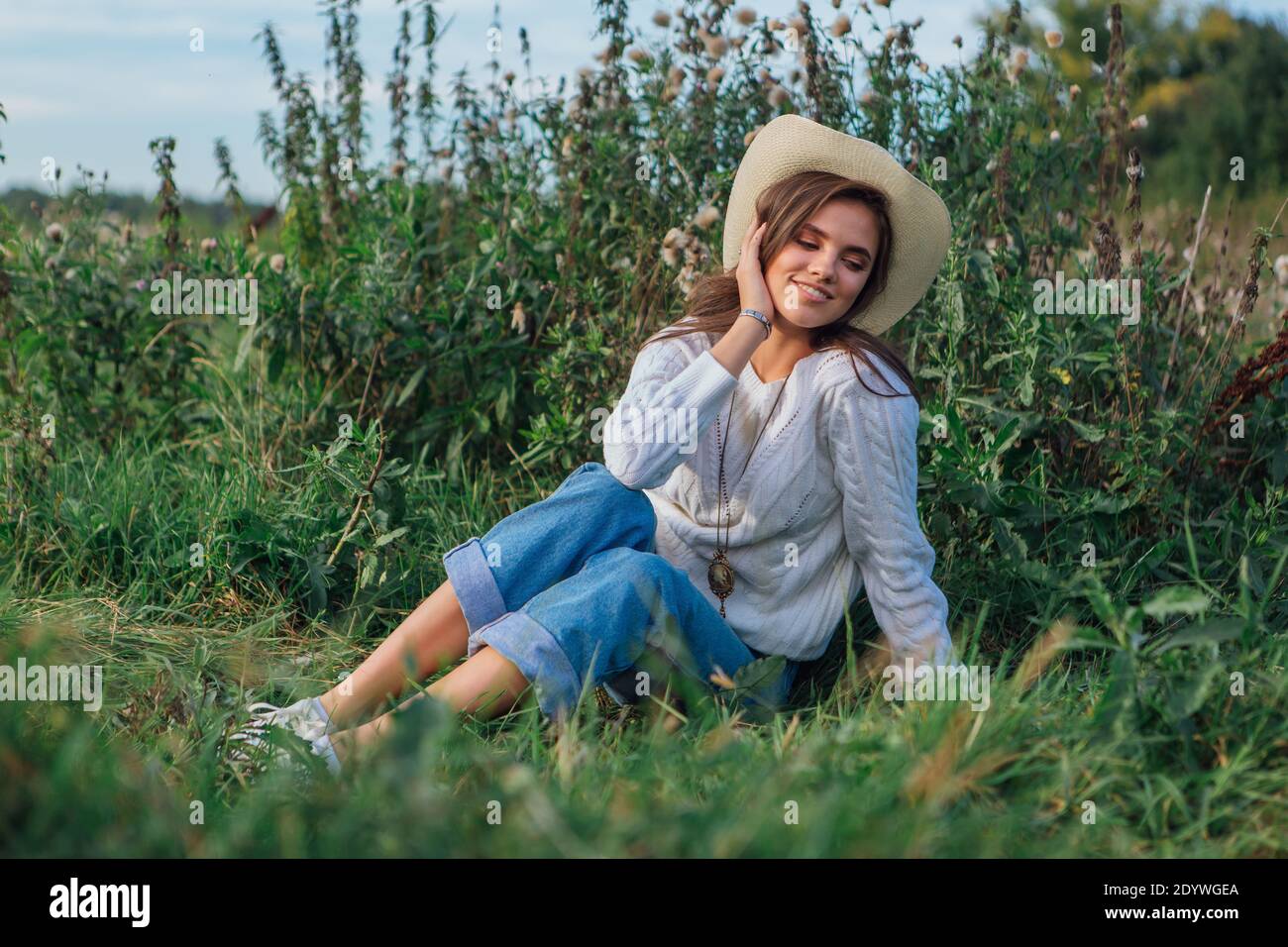 Giovane bella donna bruna vestita di un maglione bianco, jeans e cowboy cappello di paglia seduta su erba verde, sorridente e ridente durante il tramonto Foto Stock