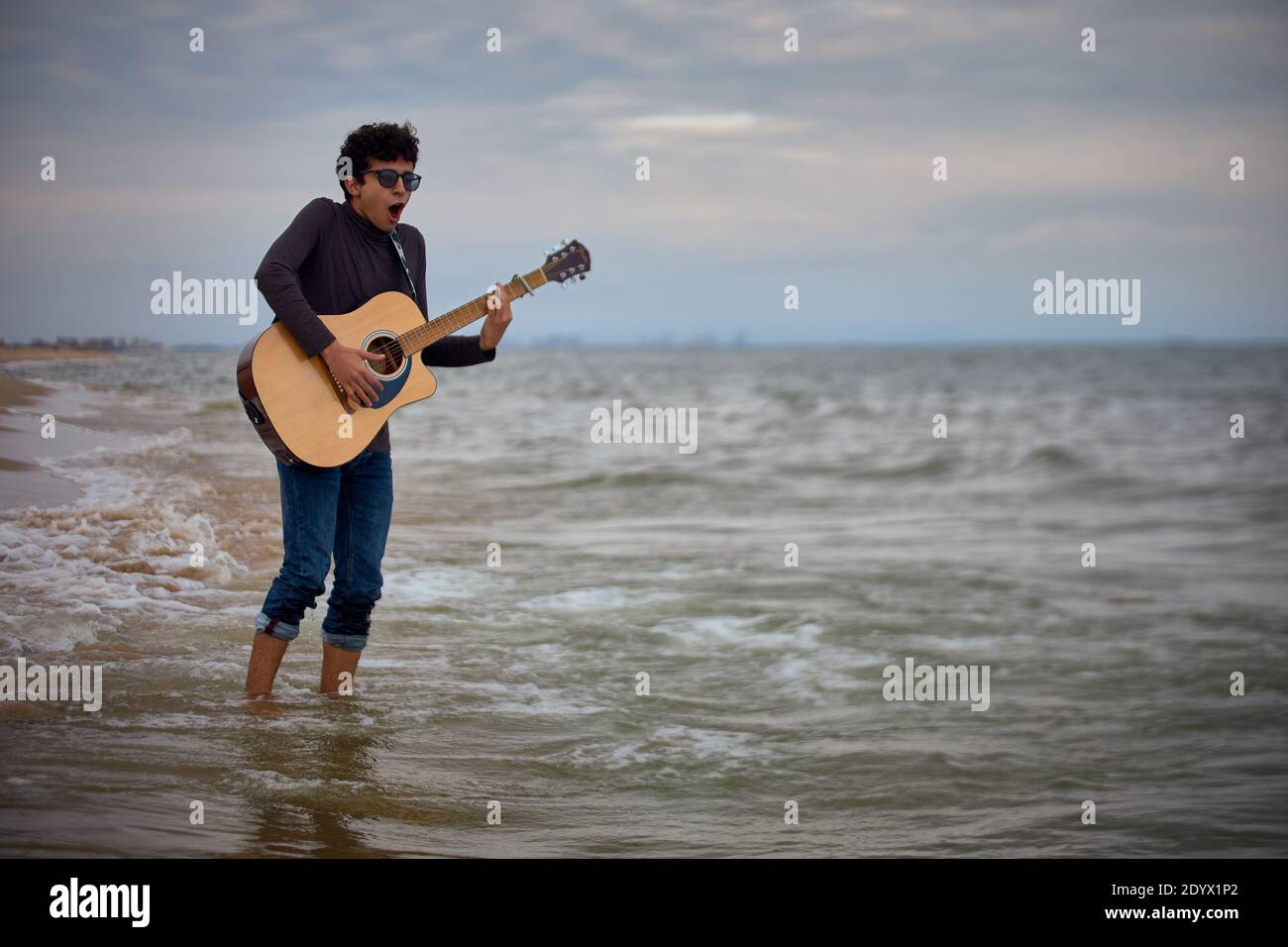 giovane adolescente caucasico che suona la chitarra acustica sulla spiaggia in acqua. occhiali da sole e vestiti scuri Foto Stock