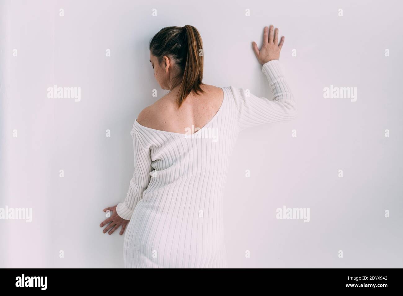 Donna che indossa un lungo abito a coste sottile a cavaliere in piedi accanto a una parete bianca con la schiena alla fotocamera Foto Stock