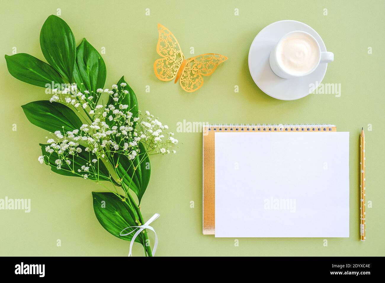 Spazio di lavoro con foglio vuoto di blocco note, matita, foglia verde, farfalla dorata decorativa e tazza di caffè su sfondo verde. Vista dall'alto piatto a T Foto Stock