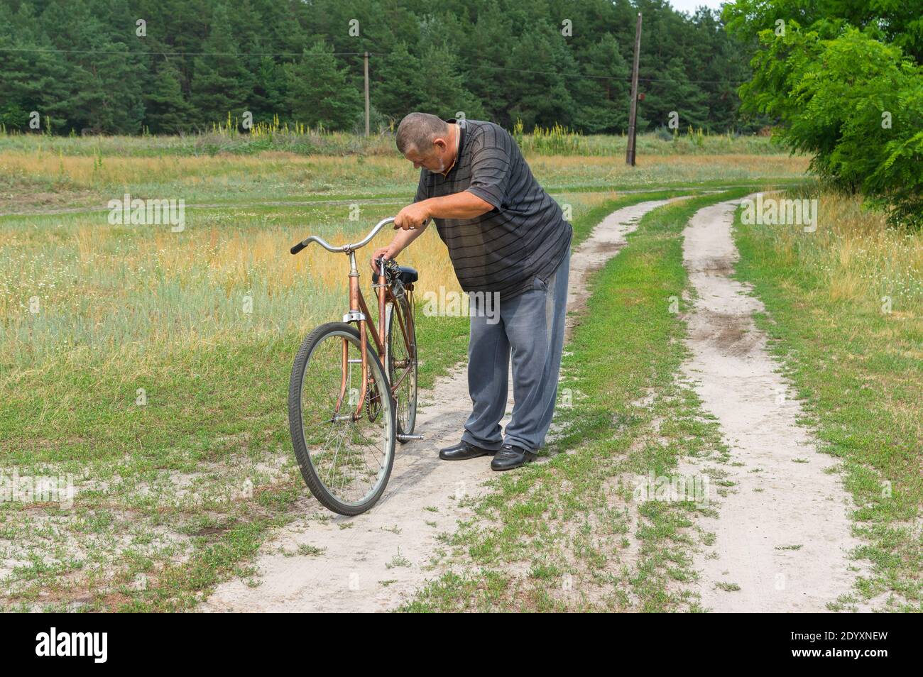 Alto contadino ucraino in piedi su una strada di campagna e ispezionando vecchia bicicletta arrugginita con sella rotta Foto Stock
