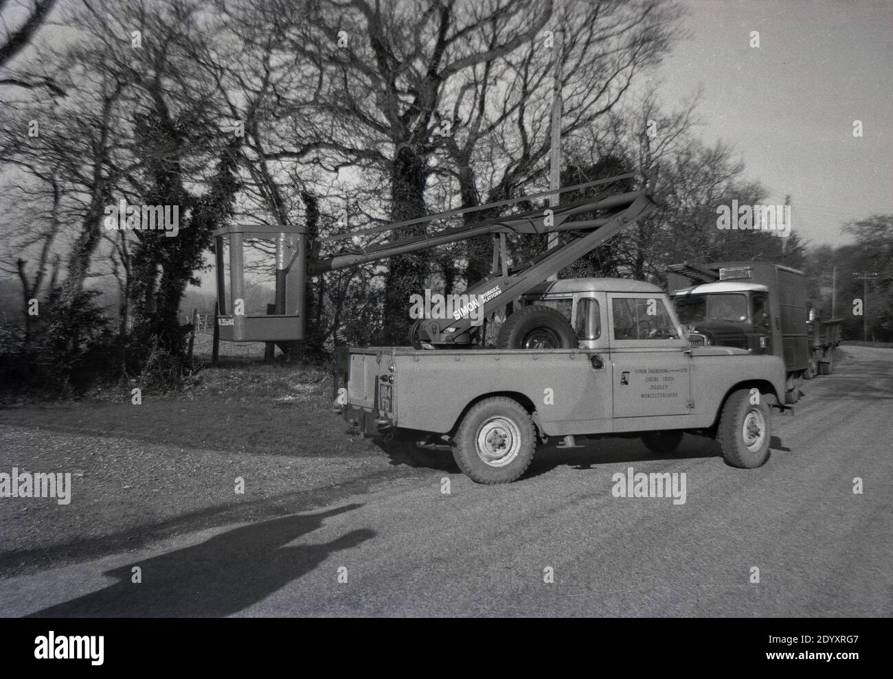 Anni '1960, storico, una piattaforma aerea sul retro di una Land rover parcheggiata su una strada di campagna, Inghilterra, Regno Unito. Tali ascensori, noti anche come raccoglitori di ciliegie o carrelli a benna, sono stati utilizzati per lavori di manutenzione e riparazione di attività stradali quali luci, cavi aerei e cavi telefonici. Foto Stock