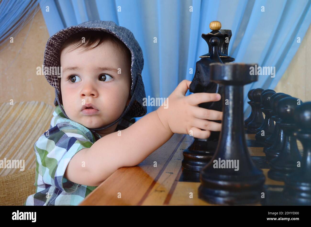 Un capretto in un cappello con gli occhi seri guarda al lato seduto un tavolo di scacchi con le figure e tenendo un cavallo con la mano. Foto Stock