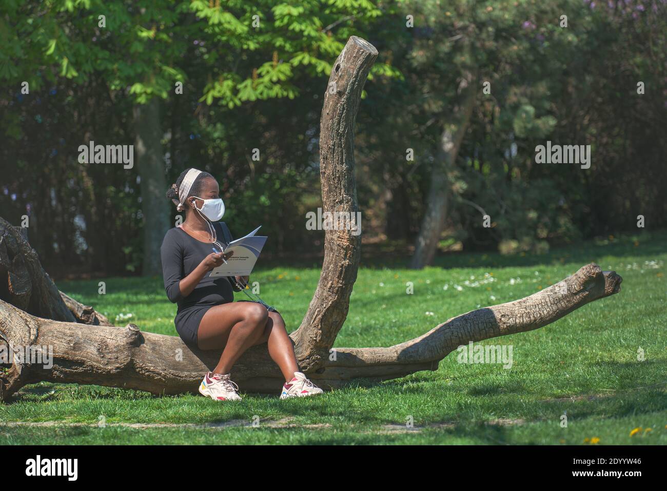 Donna afroamericana con una maschera di stoffa seduta su un tronco di albero sdraiato nel parco vicino alla coda per il test del virus corona. Foto Stock