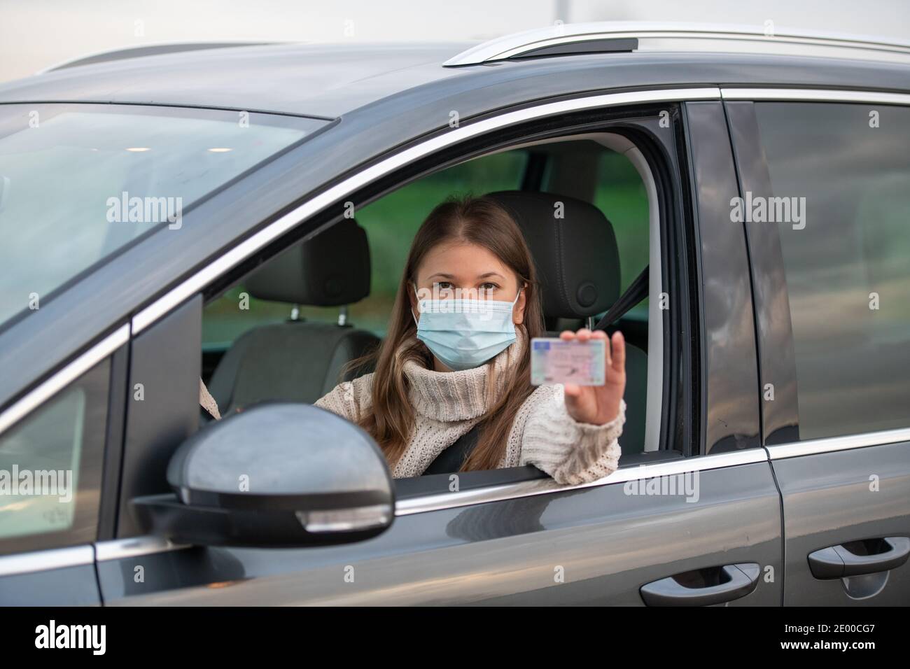 la giovane donna si siede in un'auto, indossa una maschera protettiva durante la pandemia del covid 19 e detiene la patente di guida europea Foto Stock