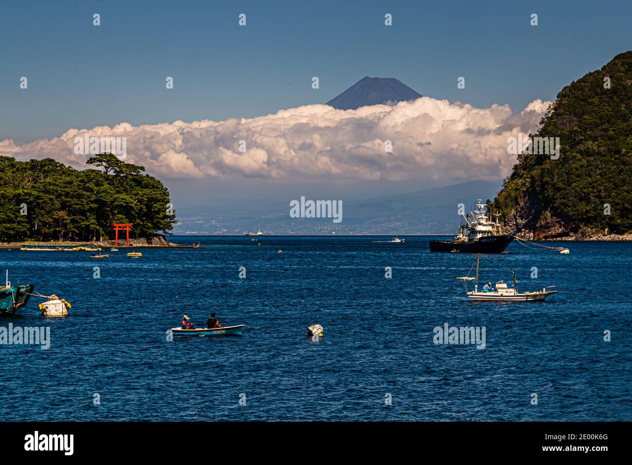 I torii rossi del Santuario di Morokuchi, protettore dei pescatori, si trovano sulla spiaggetta di Mihama-misaki nel porto di Toda; una barca da pesca e una barca a remi con due passeggeri attraversano la baia, il Monte Fuji si erge sopra le nuvole dietro di loro. Numazu, Prefettura di Shizuoka, Giappone. Foto Stock