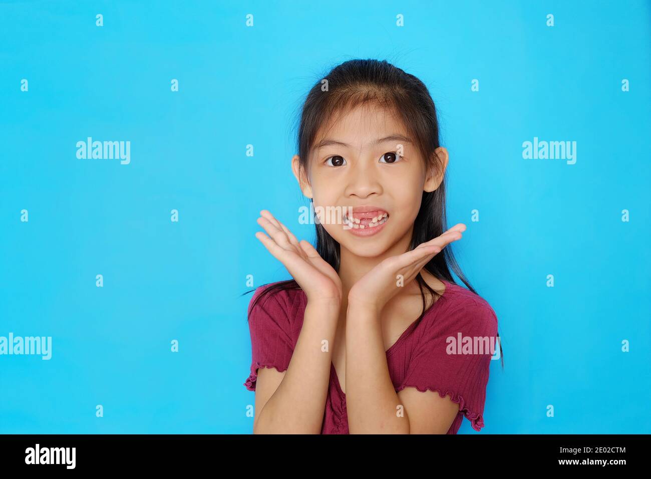 Una ragazza asiatica carina e felice con i denti anteriori mancanti, che si pone con le mani accanto al mento, con fondo azzurro chiaro e chiaro. Foto Stock