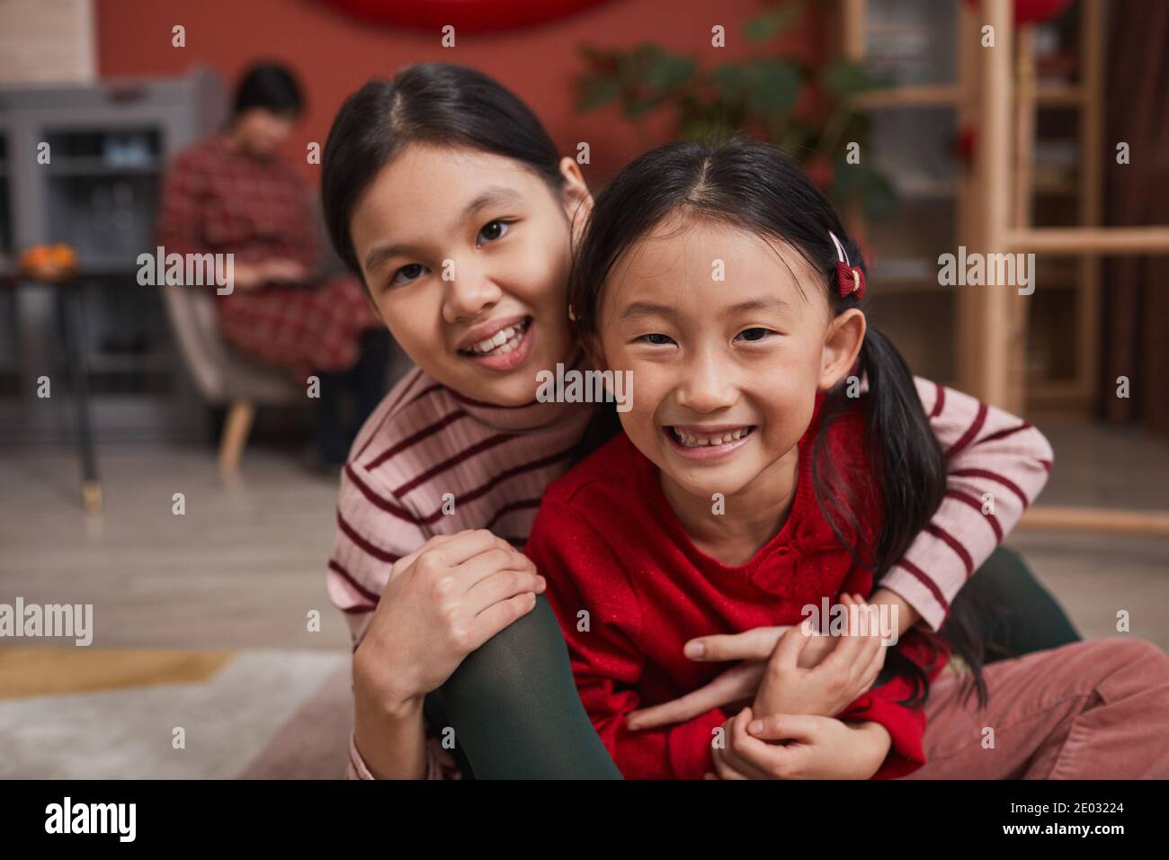 Ritratto medio ravvicinato di due giocose ragazze asiatiche che si divertono insieme in salotto sorridendo alla macchina fotografica mentre la loro madre lavorare su un computer portatile Foto Stock