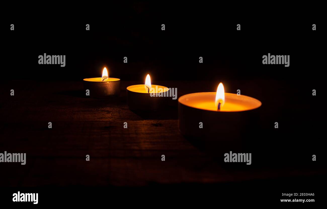 Tre candele di cera da tè illuminate su un tavolo di legno su sfondo nero. Foto Stock