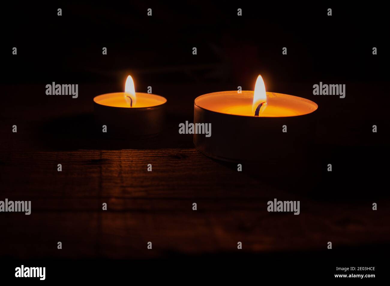 Due candele di cera da tè illuminate su un tavolo di legno su sfondo nero. Foto Stock