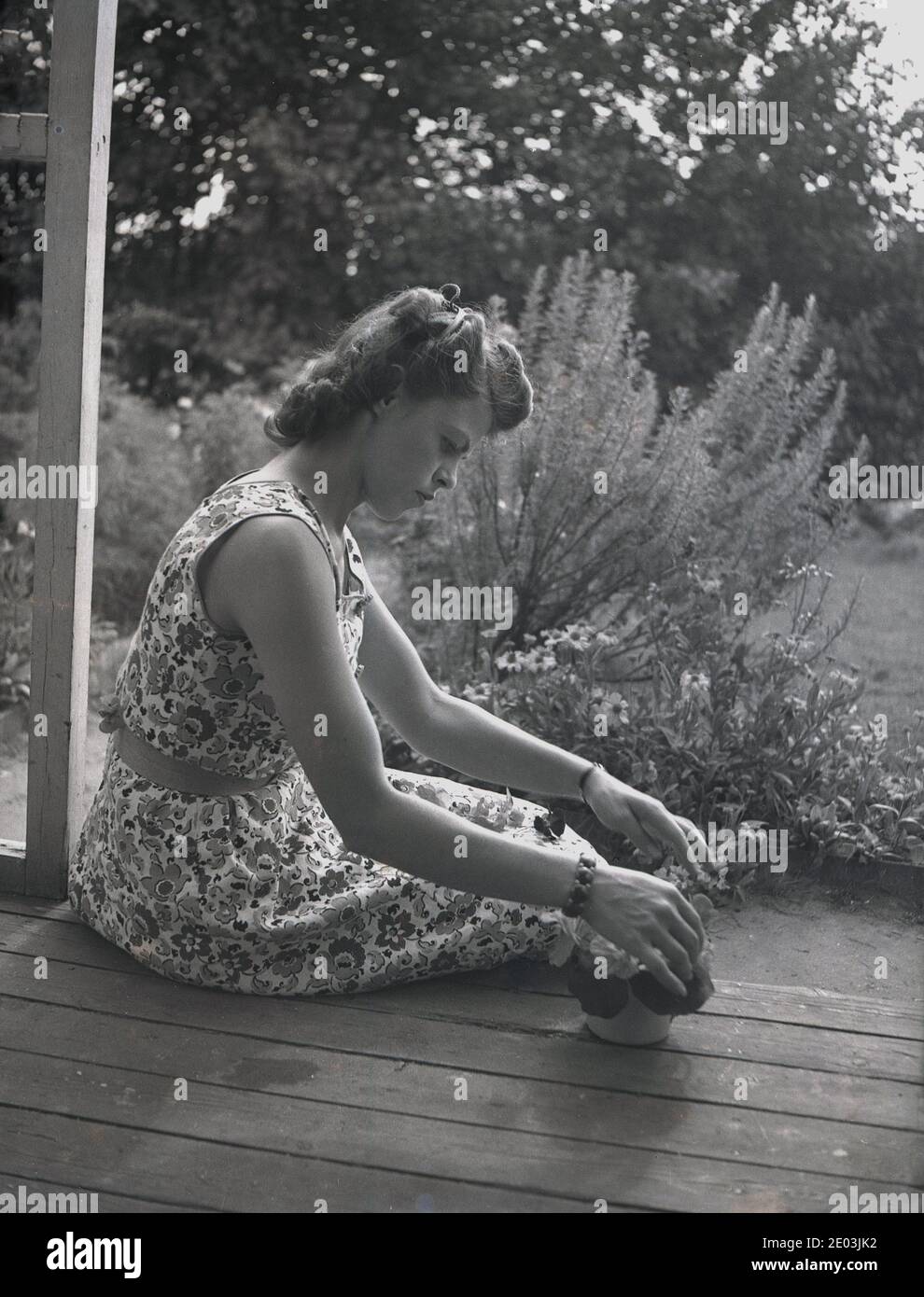 anni '1950, storica, una giovane donna che indossa un abito senza maniche a motivi floreali, un popolare oggetto di moda femminile in quest'epoca, seduto su un'area di legno sul retro di una casa vicino al giardino, sistemando i fiori in una piccola pentola. Foto Stock