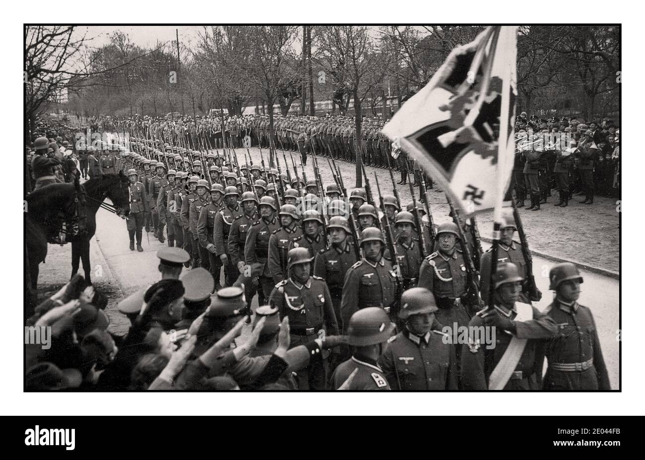 1940 Nazi Wehrmacht Army Marching Parade, con regimental bandiera standard davanti. Banda militare che suona dietro. Spettatori che danno saluto a Heil Hitler in primo piano. Foto Stock
