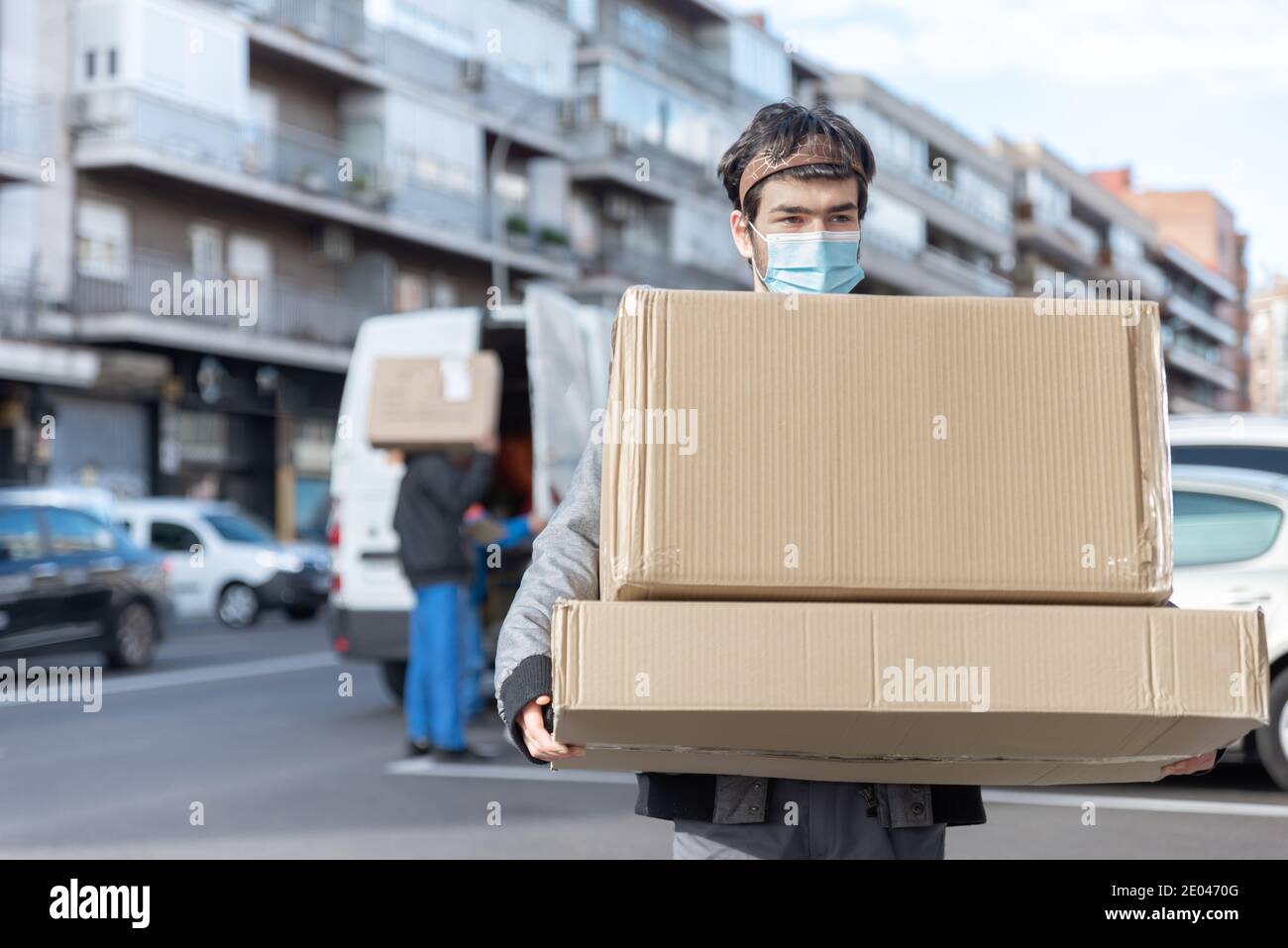 L'uomo di consegna con uniforme e maschera porta una grande consegna box e furgone fuori fuoco in background Foto Stock