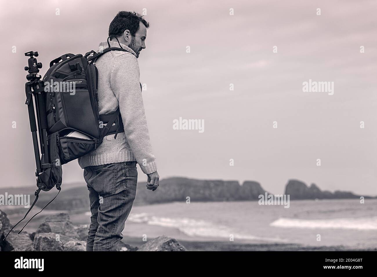 Fotografo irlandese su una spiaggia sulla costa di rame dell'Irlanda. Con un grande zaino e un treppiede sulla schiena, guardando il mare. Foto Stock