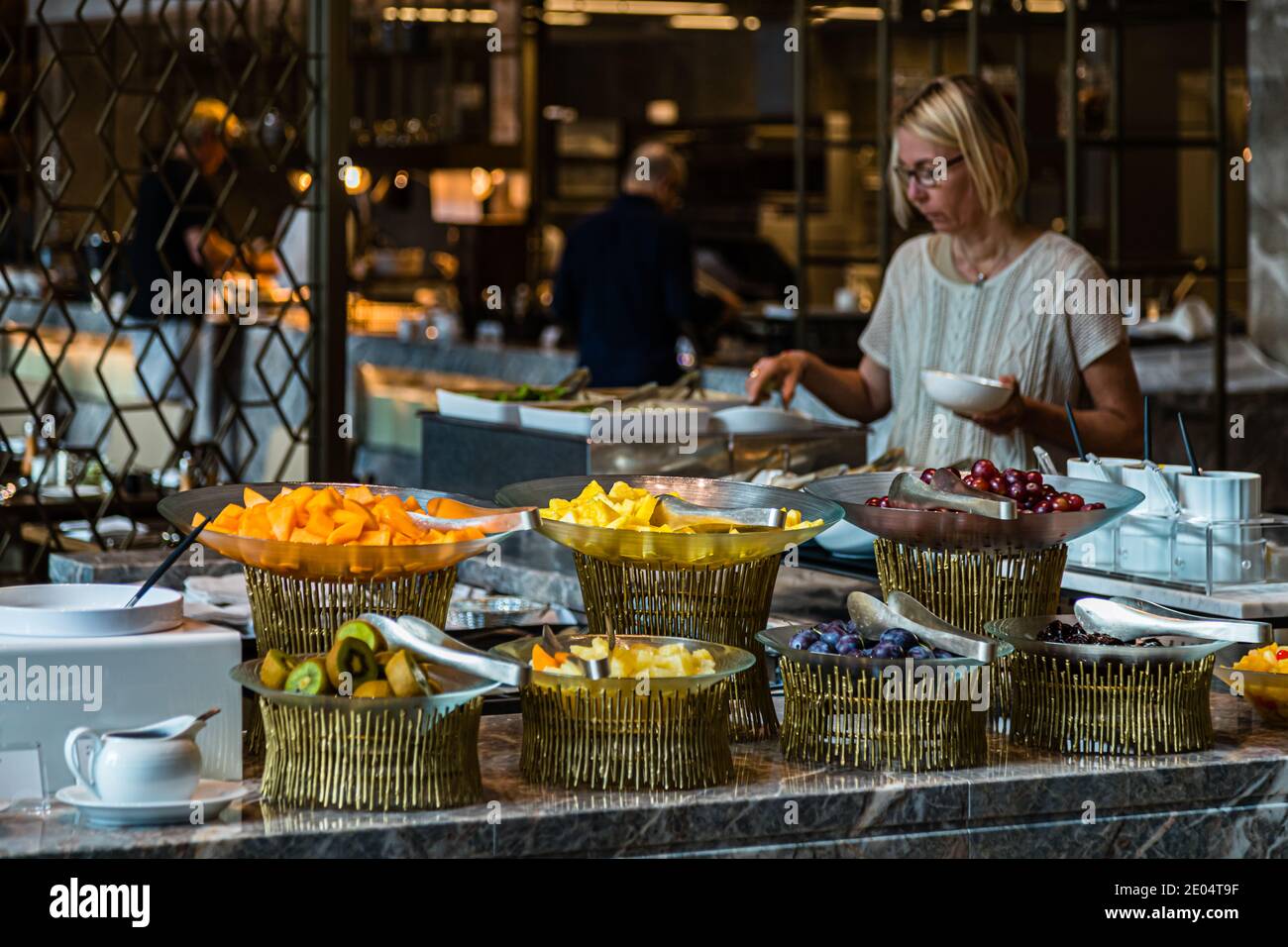 Cestini dorati di frutta fresca – mango, ananas, kiwi, uva – riempiono il banco del buffet della colazione all’Okura Tokyo, nel quartiere di Minato, Giappone. Un ospite sceglie la frutta mentre il personale prepara il cibo sullo sfondo. L’hotel di lusso ha riaperto nel settembre 2019 dopo una ristrutturazione di quattro anni. Foto Stock