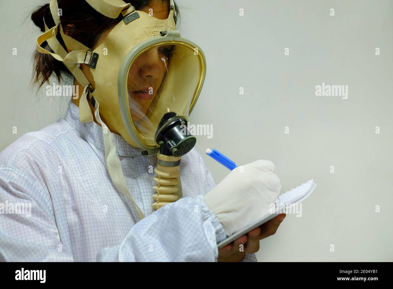 Una donna con una maschera protettiva per l'ossigeno a pieno viso, camice bianco e guanti in gomma, pronta a lavorare in un laboratorio biochimico, prendendo appunti con la penna A. Foto Stock