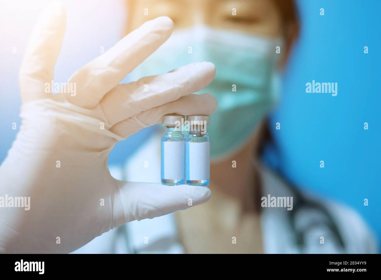 Una donna asiatica con maschera chirurgica e guanti di gomma bianca in una clinica, che contiene due bottiglie di vetro identiche di vaccino con etichetta bianca vuota Foto Stock