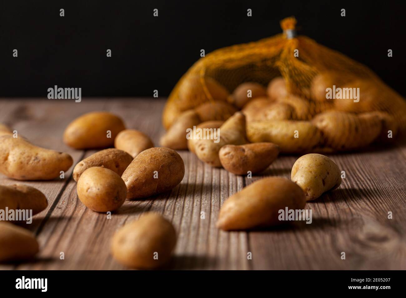 Profondità di campo poco profonda immagine scura di piccole patate ratte di forma irregolare sparse sulla superficie di un tavolo di legno da un sacco di maglia rovesciato. An Foto Stock