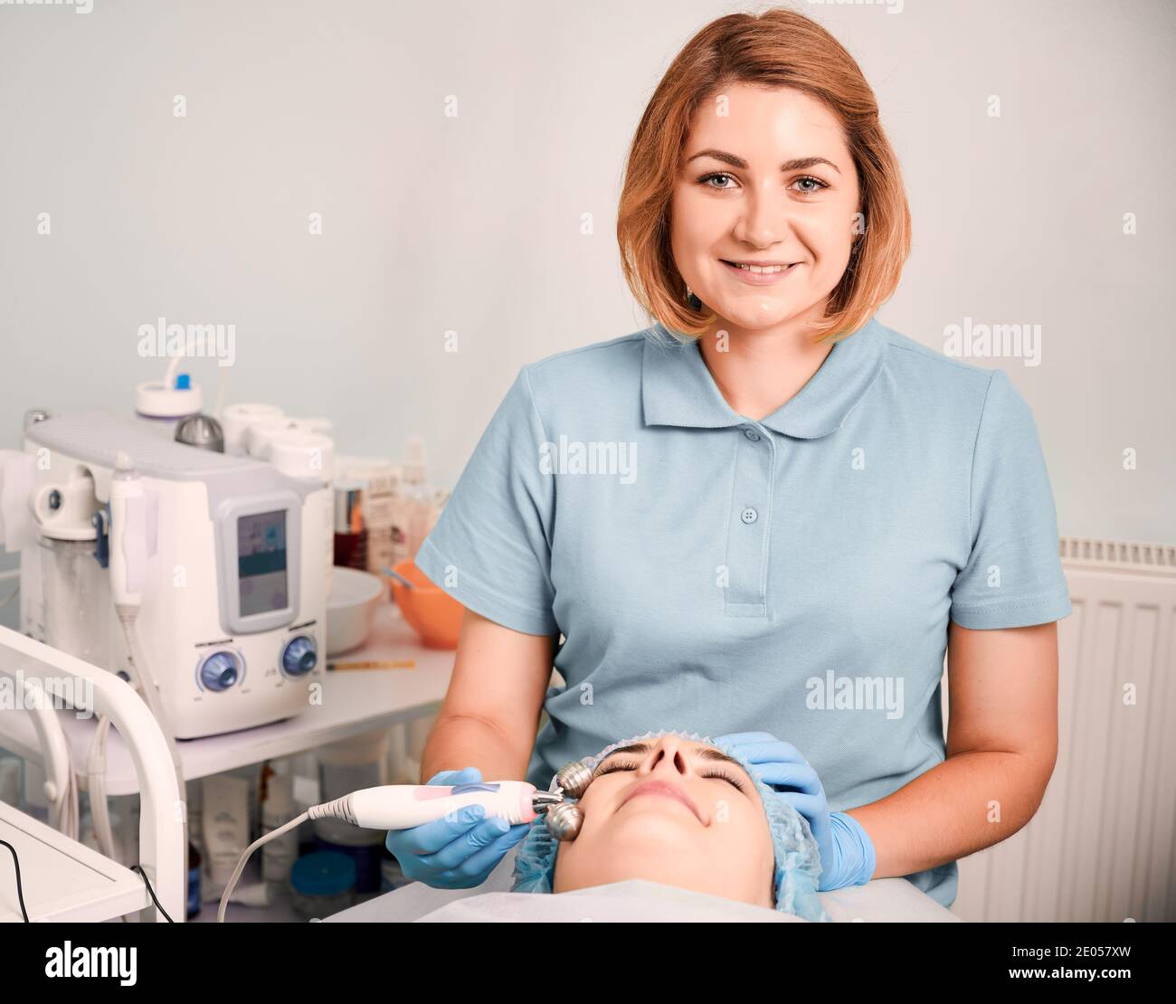 Cosmetologa massaggiare il viso femminile del cliente con un rullo anti-invecchiamento per il viso in microcorrente. Donna in cappello monouso sdraiata sul letto da giorno mentre si ha un massaggio galvanico al centro benessere. Concetto di bellezza. Foto Stock