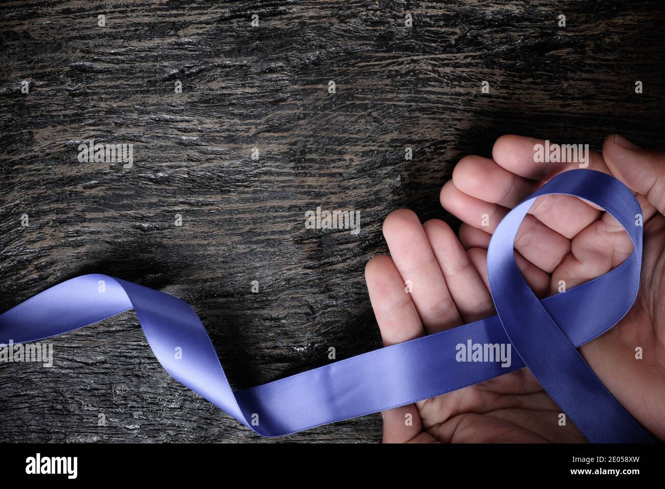 Vista dall'alto della mano maschio che tiene il nastro blu perivinkle su sfondo di legno con spazio di copia. Consapevolezza del cancro dello stomaco e dell'esofago, disturbi alimentari Foto Stock