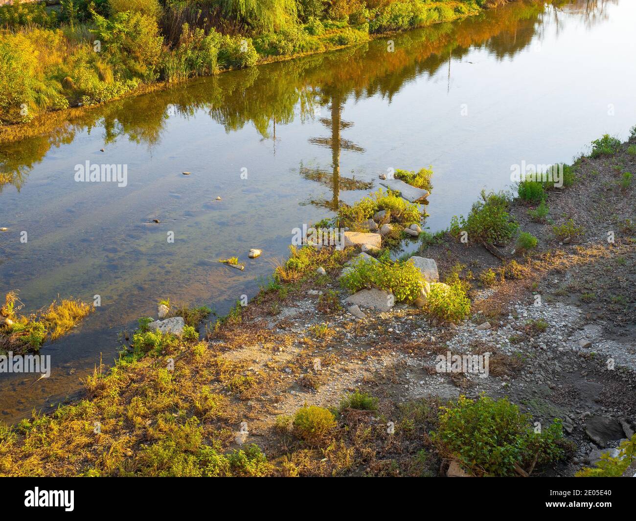 Un fiume con acqua calma e un fondo fangoso riflette il cielo e fitti spessi di erba ingiallente sul shore Foto Stock