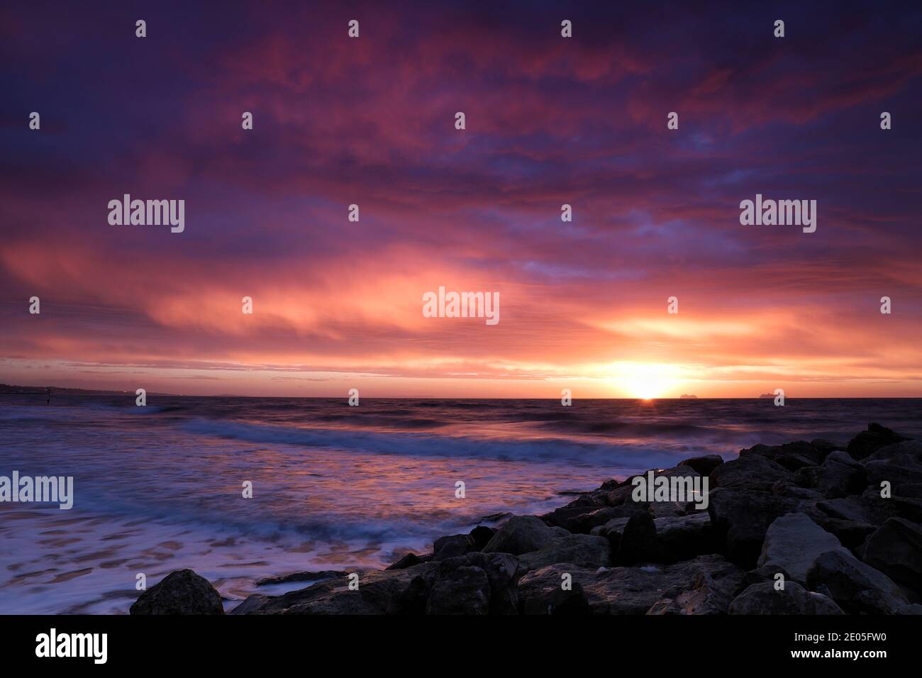 Il sole che sorge riempie un cielo pieno di nubi canore da sogno con magici colori rosa e viola, mentre le onde si infrangono sulla riva in un caos gelido. Foto Stock