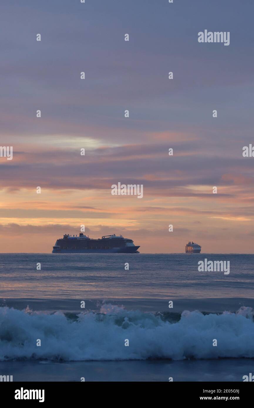 Un paio di navi da crociera può essere visto all'orizzonte sotto un cielo pieno di nuvole color pastello mentre le onde rotolano verso la riva in una schiuma bianca. Foto Stock