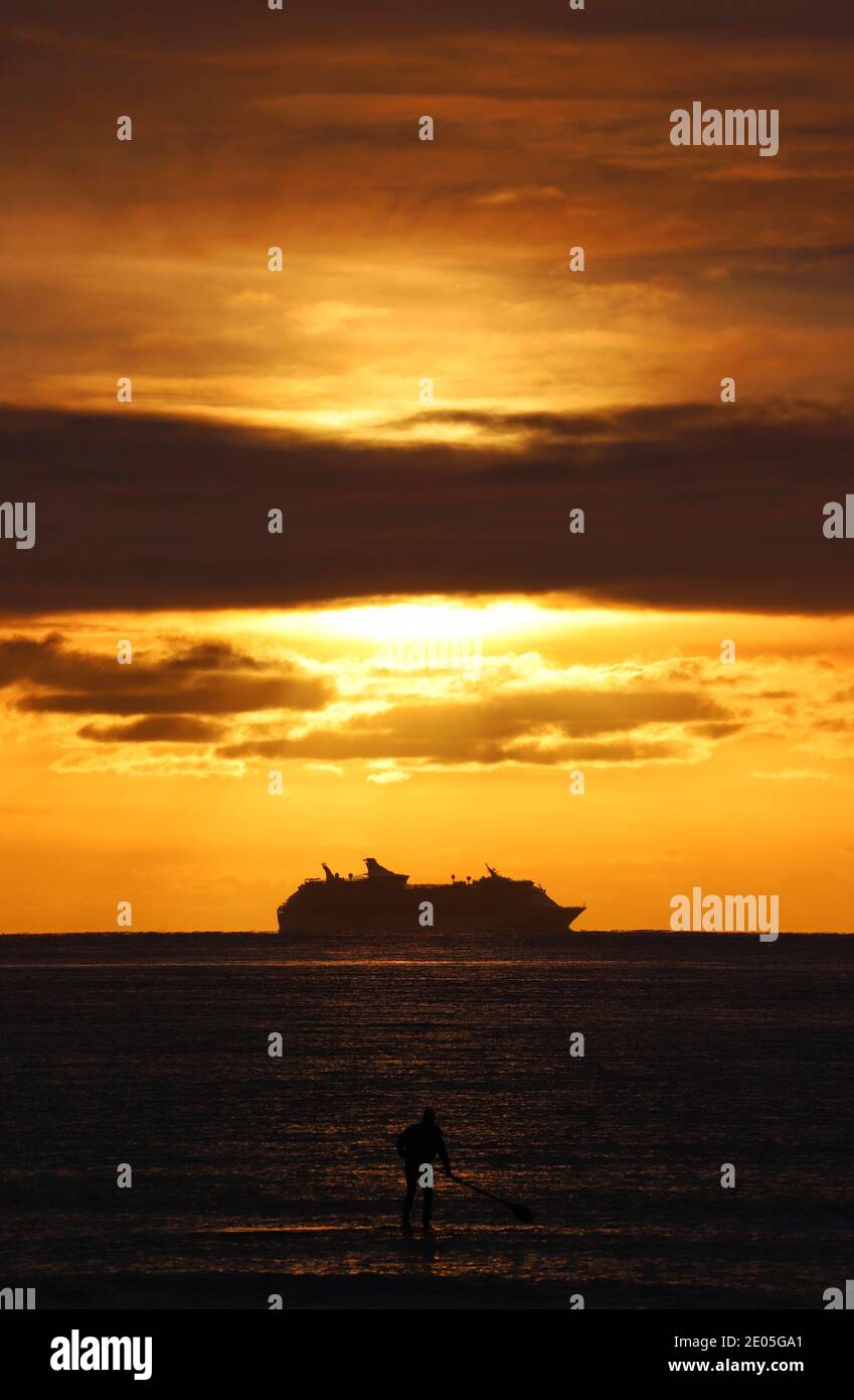 Una grande nave da crociera può essere visto scivolare sopra l'orizzonte sotto la luce dorata del sole nascente mentre un boarder di pagaia è silhoueted sul mare. Foto Stock