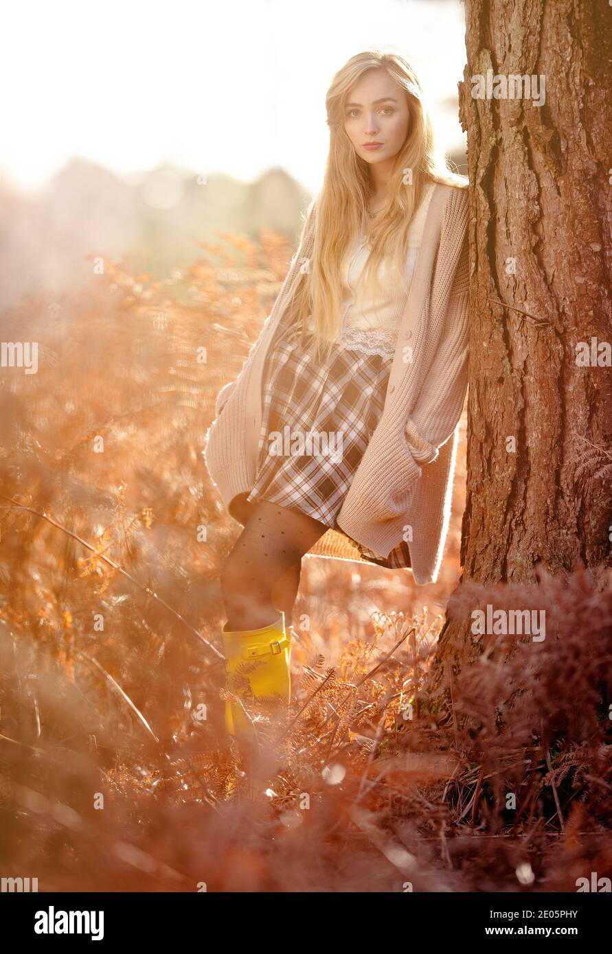 Una giovane bella donna (20) Dreamy editoriale di moda girato dentro una calda foresta autunnale con cardigan e tartan gonna con lunghi capelli biondi Foto Stock