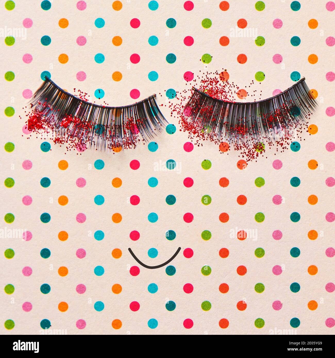 un paio di ciglia finte cosparse di glitter rosso e. una bocca sorridente che forma un volto felice su una pallida sfondo rosa modellato con punti di differen Foto Stock