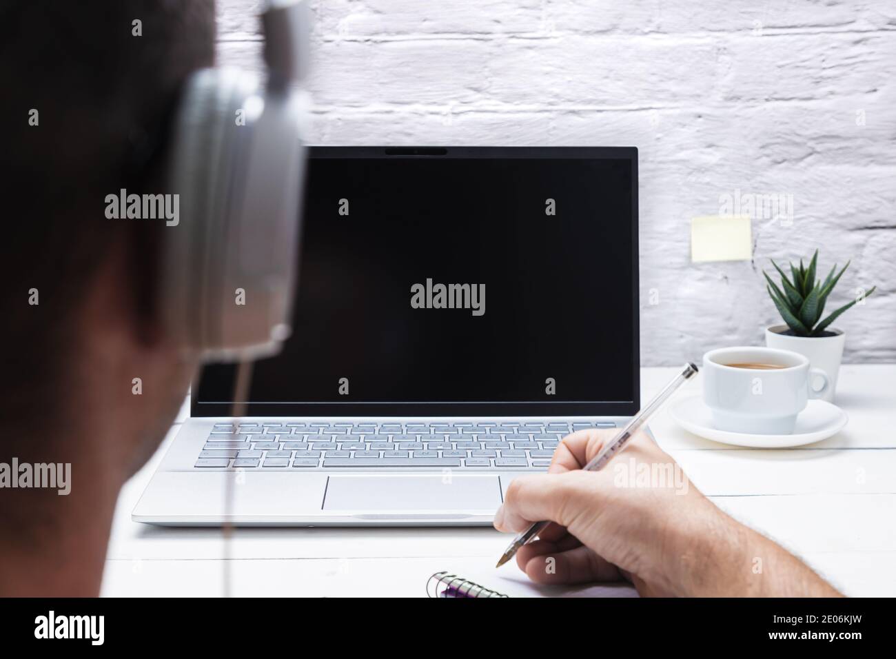 Uomo sulla schiena con le cuffie che prendono appunti guardando il computer portatile e bevendo caffè su sfondo bianco. Foto Stock