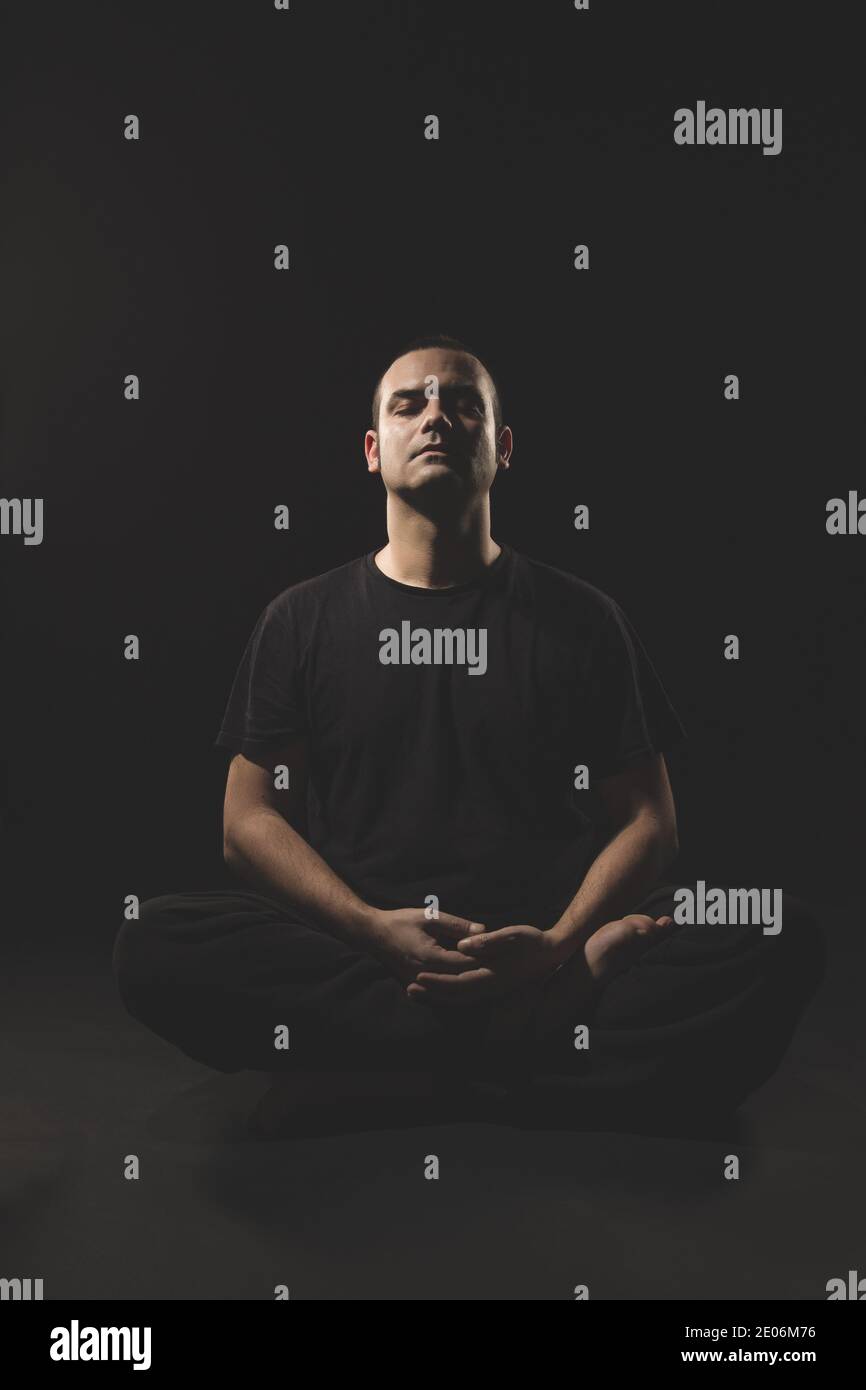 Giovane caucasico seduto in meditazione con vestiti neri e sfondo nero. Foto Stock