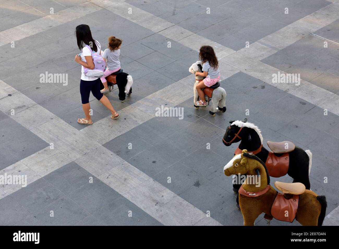 Bambini che cavalcano su cavalli giocattolo a piedi nella piazza pubblica Foto Stock