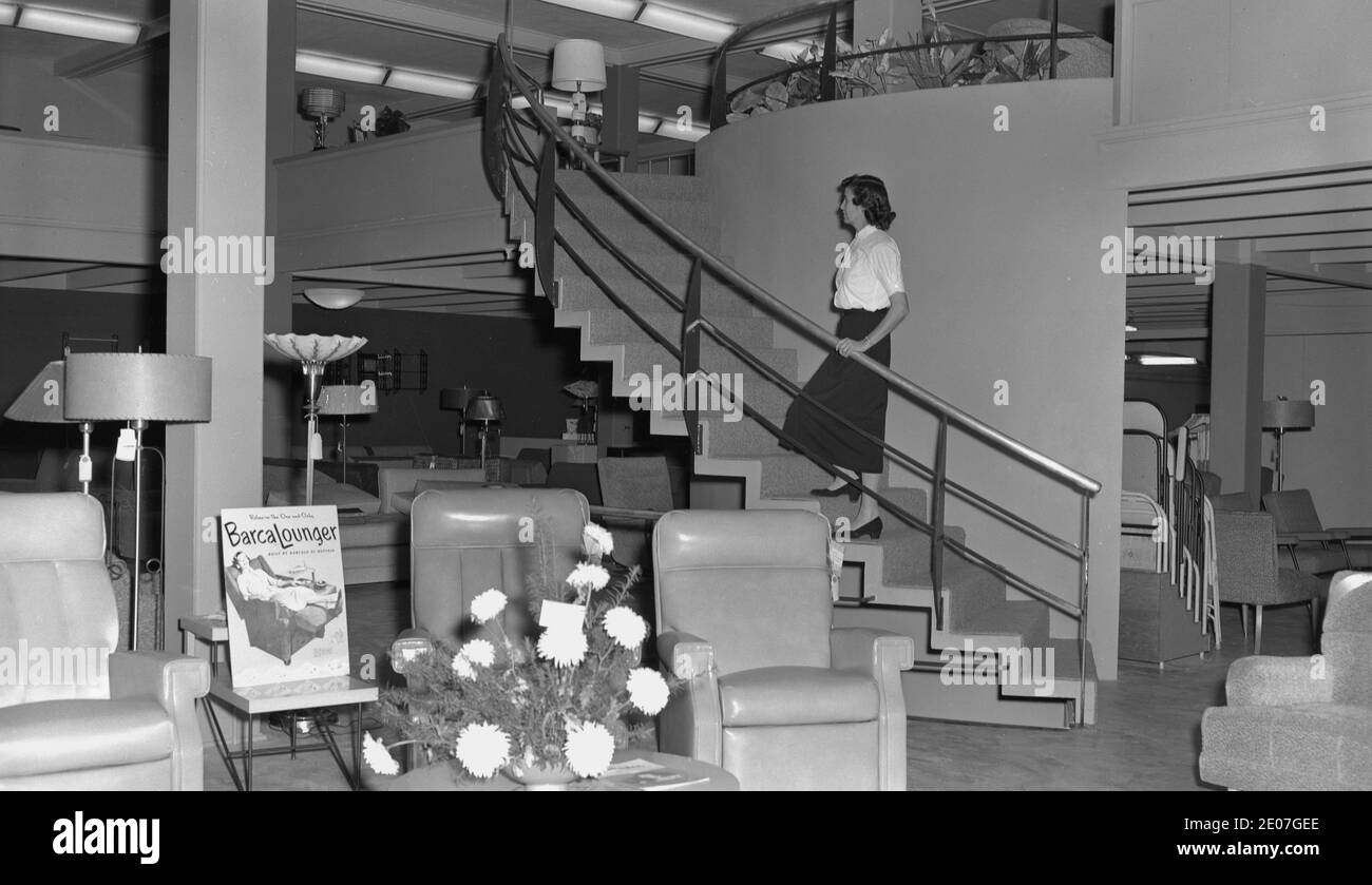 Anni '1950, storica, una signora sulle scale in un negozio di mobili, USA. Su una delle sedie in mostra, un annuncio per una Barcalounger, una sedia reclinabile, realizzata dalla Barcalounger Company. Il più antico produttore di sedie reclinabili in America, l'azienda è stata fondata nel 1896 a Buffalo, NewYork, come Barcalo Manufacturing Company, dopo che il suo fondatore, Edward J. Barcalo, aveva originariamente realizzato utensili, prima di trasferirsi nei mobili. Foto Stock